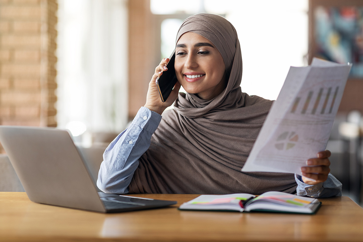 Muslim business woman working at cafe, having business conversation on mobile phone, holding marketing research and looking at laptop screen, copy space